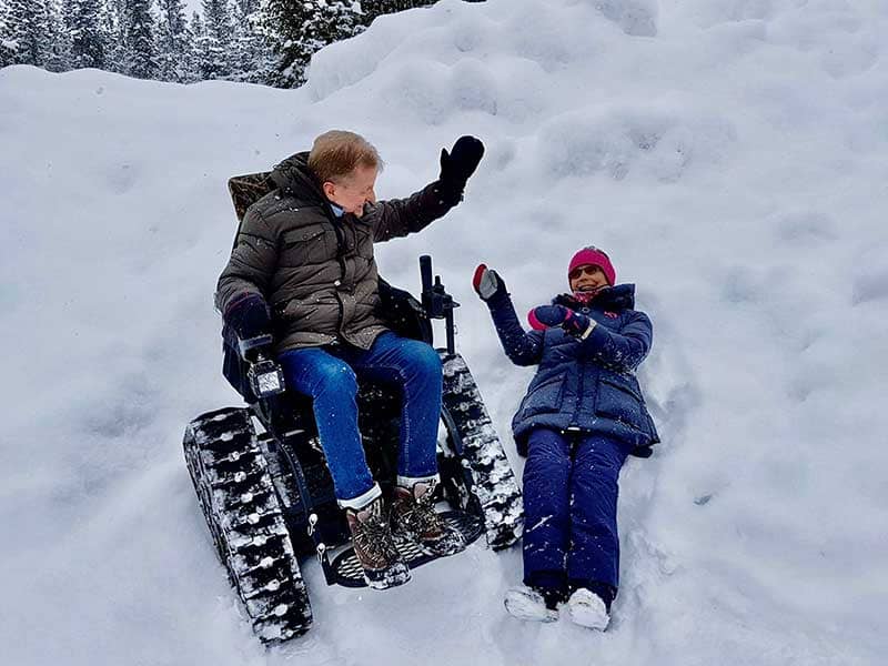 Sven-Erik sitter i Maries Action trackchair på fjället och Marie ligger i snön och skrattar